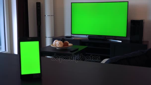 Um smartphone em uma mesa em uma posição vertical uma TV em segundo plano, ambos têm telas verdes — Vídeo de Stock