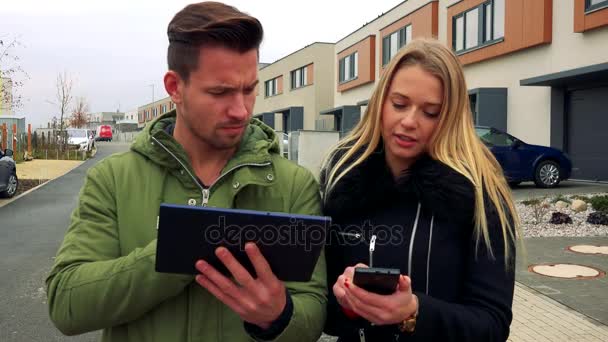 Ένας άνδρας και μια γυναίκα συζητούν κάτι σχετικά με ένα δισκίο και ένα smartphone σε ένα δρόμο σε μια γειτονιά — Αρχείο Βίντεο