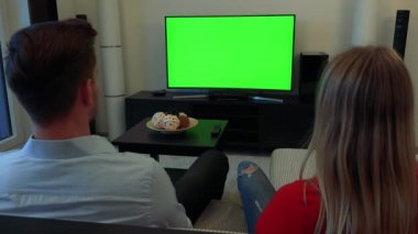 Bir erkek ve bir kadın (başlarını sırt kameraya) rahat bir oturma odasında yeşil bir ekran bir Tv izle