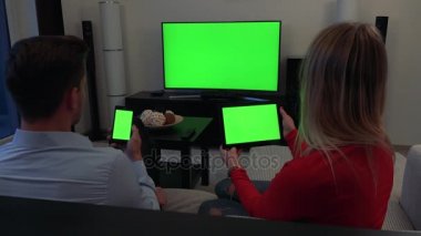 Bir erkek ve bir kadın kanepede oturup, o bir akıllı telefon tutar, o tüm üç yeşil ekran tablet, bir Tv içinde belgili tanımlık geçmiş - tutar