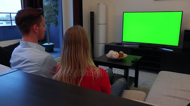 Молодая, привлекательная пара смотрит телевизор с зеленым экраном в уютной гостиной, затем поворачивается к камере и улыбается — стоковое видео