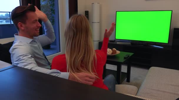 Een paar jonge, aantrekkelijke horloges van een Tv met een groen scherm in een gezellige huiskamer en viert — Stockvideo