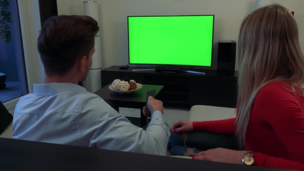 Молода, приваблива пара дивиться телевізор із зеленим екраном у вітальні, чоловік перемикається між каналами, жінка тремтить головою і бере на себе контролер — стокове відео