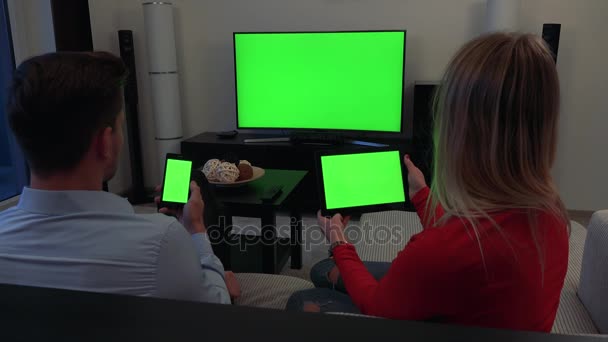 Um homem e uma mulher sentam-se em um sofá, ele segura um smartphone, ela segura um tablet, uma TV no fundo - todas as três telas verdes — Vídeo de Stock