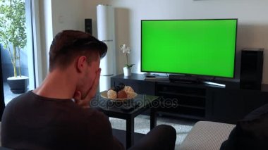 Bir adam rahat bir oturma odasında yeşil bir ekran bir Tv izliyor ve üzgün ve hayal kırıklığı onun kollarında yüzünü gömer