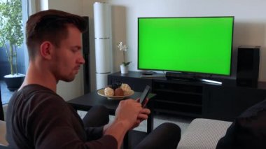 Genç ve yakışıklı bir adam rahat bir salon ve türleri yeşil bir ekran bir Tv onun smartphone üzerinde saatler.