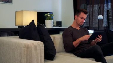 Genç ve yakışıklı bir adam rahat bir oturma odasında bir kanepede oturuyor ve onun tablet kullanır