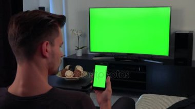 Kafasının arkasını kameraya ile açık bir adam bir Tv izliyor ve sonunda elinde bir smartphone vasıl bakışları, Tv ve telefon yeşil bir ekran var