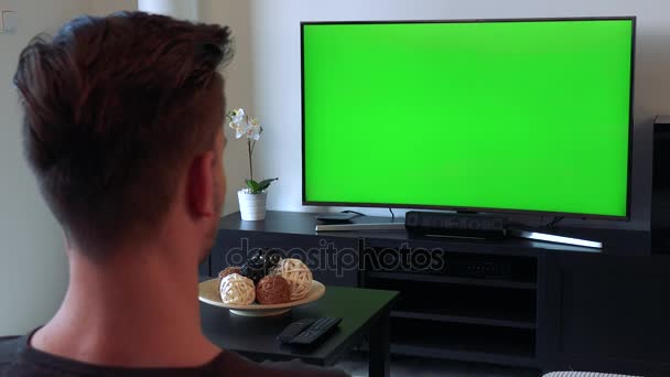 Un uomo, dietro la testa alla telecamera, guarda una TV con uno schermo verde in una stanza accogliente — Video Stock