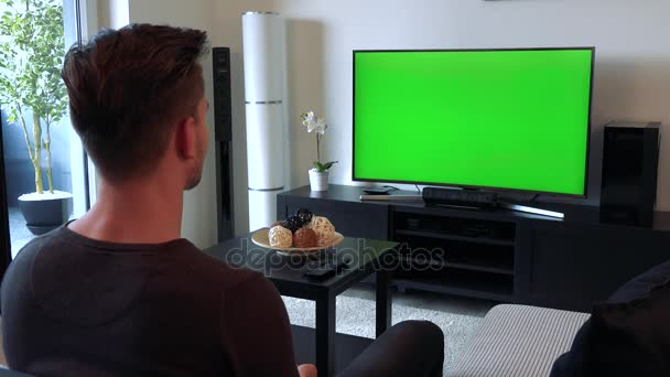 Молодой, красивый мужчина смотрит телевизор с зеленым экраном, затем поворачивается к камере и улыбается — стоковое видео