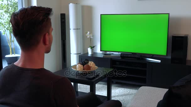 Молодой, красивый мужчина смотрит телевизор с зеленым экраном, затем поворачивается к камере, улыбается и показывает большой палец вверх — стоковое видео