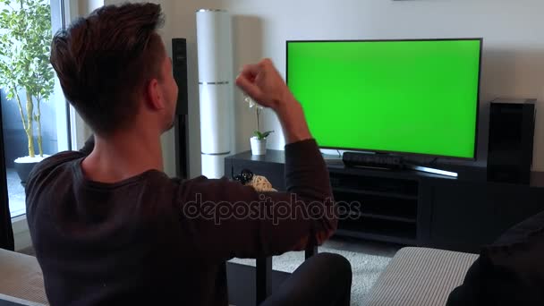 Um homem assiste uma TV com uma tela verde em uma sala de estar aconchegante e começa a comemorar (gritar alegremente, bater palmas e levantar triunfalmente os punhos ) — Vídeo de Stock