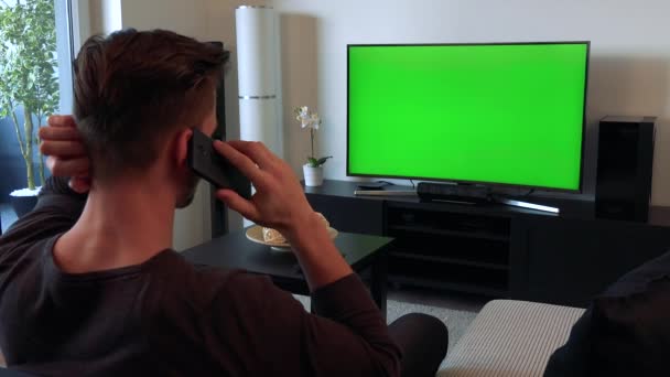 Een man praat op een smartphone en een Tv met een groen scherm horloges — Stockvideo