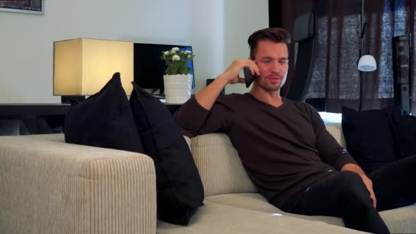 Un hombre joven y guapo habla en un smartphone y se sienta en un sofá — Vídeos de Stock