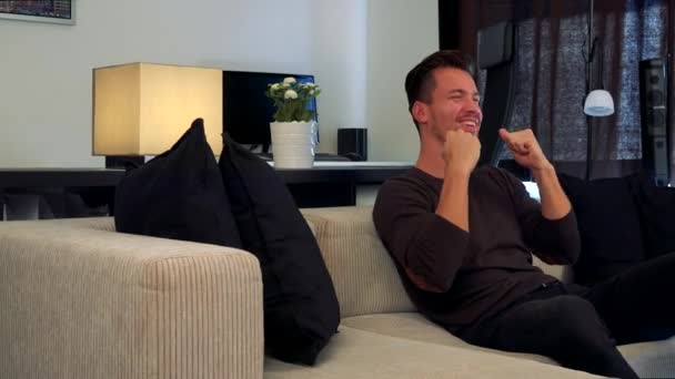 A young, handsome man sits on a couch and celebrates — Stock Video