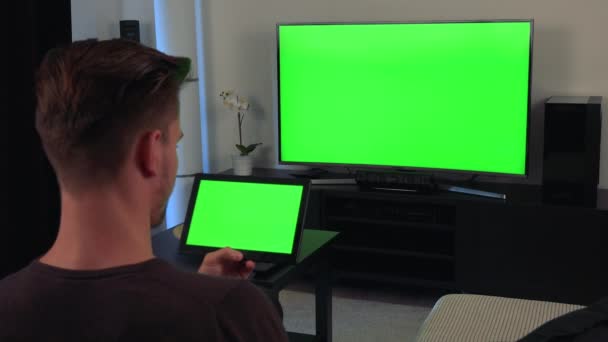 En man, baksidan av huvudet vände till kameran, klockor en anteckningsbok och en Tv, båda med grön skärm — Stockvideo
