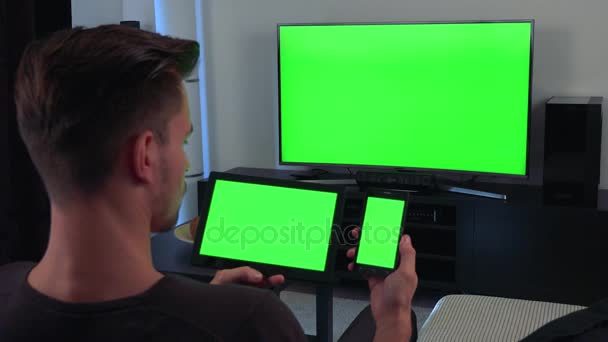 A man, the back of his head turned to the camera, holds a smartphone, a tablet and a TV, all three with green screens — Stock Video