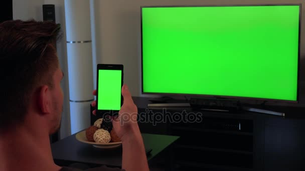 Мужчина, повернутый затылком к камере, смотрит телевизор и держит смартфон, оба с зеленым экраном — стоковое видео