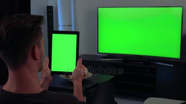 En man, baksidan av huvudet vände till kameran, innehar en tablett och klockor Tv, båda med en grön skärm — Stockvideo