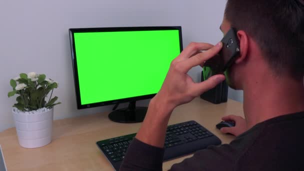 Un uomo parla su uno smartphone mentre su un computer con uno schermo verde — Video Stock