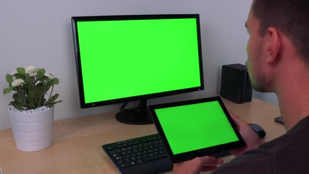 Ένας άνθρωπος ρολόγια στις στροφές του tablet και μια οθόνη υπολογιστή, δύο πράσινες οθόνες — Αρχείο Βίντεο