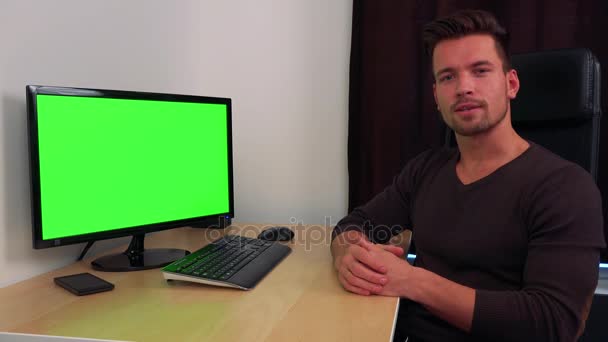 Um jovem e bonito homem se senta em uma mesa na frente de uma tela de computador verde e explica algo para a câmera — Vídeo de Stock