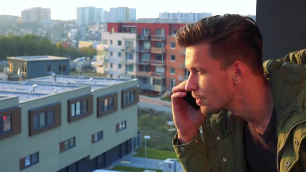 Un hombre joven y guapo habla en un smartphone en un balcón — Vídeos de Stock