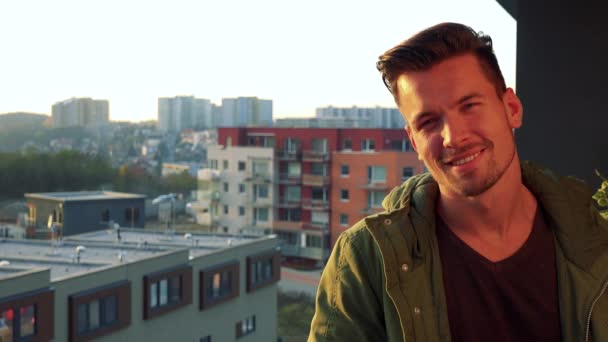 Een jonge, knappe man staat op een balkon, kijkt uit en glimlacht op de camera — Stockvideo