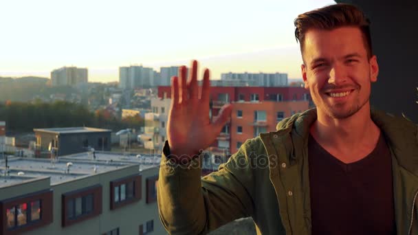 Een jonge, knappe man staat op een balkon, glimlacht en golven op de camera — Stockvideo