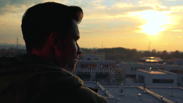 A man, the back of his head to the camera, looks around at the city below him during a sunset — Stock Video