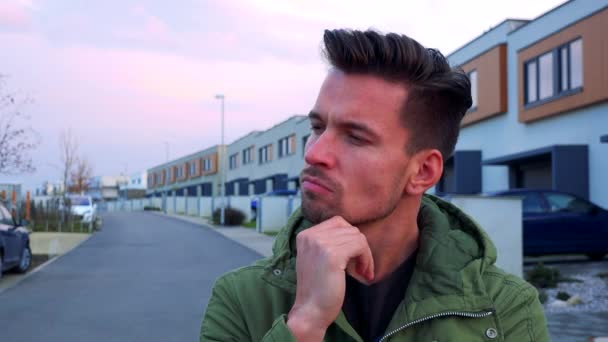 Ein junger, gutaussehender Mann steht auf einer Straße vor der Kamera und denkt hart über etwas nach, Häuser im Hintergrund — Stockvideo