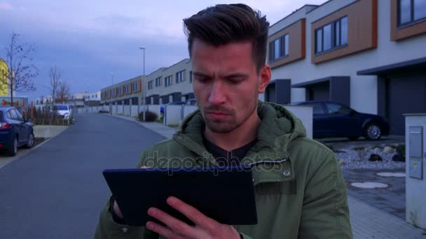 Een jonge, knappe man staat op een weg in een buurt en werkt op een Tablet PC — Stockvideo