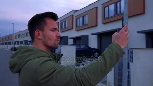 En ung, stilig man står på en väg i ett område och tar en bild på en smartphone — Stockvideo