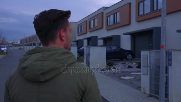 Ένα νεαρό, όμορφος άντρας στέκεται με την πλάτη στην κάμερα σε ένα δρόμο σε μια γειτονιά και κοιτάζει γύρω — Αρχείο Βίντεο