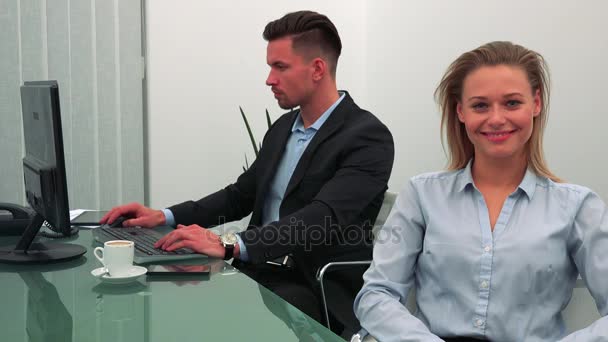 Egy fiatal, gyönyörű nő ül egy asztal, egy iroda, és mosolyog a kamera, kollégája szerkezet-ra egy számítógép, a háttérben — Stock videók