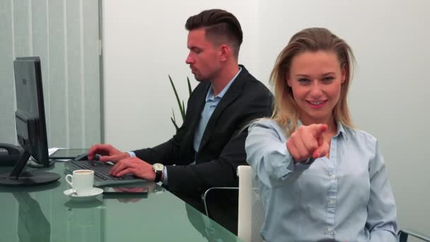 A young, attractive woman smiles and points at the camera in an office, her colleague works on a computer in the background — Stock Video