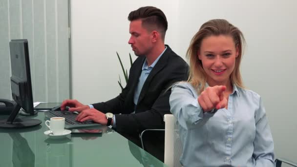 Mladá, atraktivní žena usměje a ukazuje na kameru v kanceláři, její kolega práce na počítači na pozadí