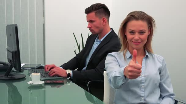 A young, attractive woman smiles and shows a thumb up to the camera in an office, her colleague works on a computer in the background — Stock Video