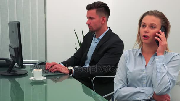 Μια νεαρή, όμορφη γυναίκα συνομιλίες σε ένα smartphone σε ένα γραφείο, τον συνάδελφό της που λειτουργεί σε έναν υπολογιστή στο παρασκήνιο — Αρχείο Βίντεο