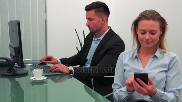 Een jonge, aantrekkelijke vrouw werkt op een smartphone in een kantoor, haar collega werkt op een computer op de achtergrond — Stockvideo