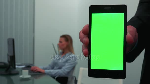 Смартфон с зеленым экраном - крупным планом, деловая женщина работает за компьютером на заднем плане — стоковое видео