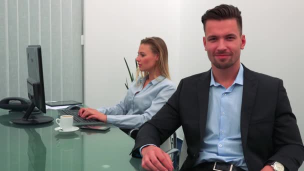 Een jonge, knappe man achter een bureau in een kantoor en glimlacht op de camera zit, zijn collega werkt op een computer op de achtergrond — Stockvideo