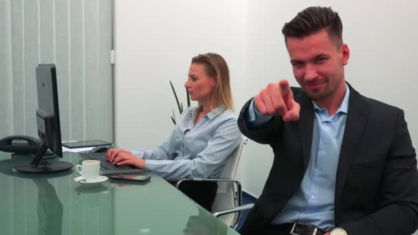 Um homem jovem e bonito sorri e aponta para a câmera em um escritório, seu colega trabalha em um computador ao fundo — Vídeo de Stock