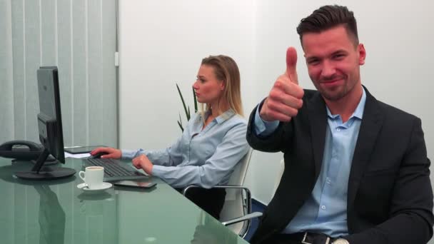 Un hombre joven y guapo sonríe y muestra un pulgar hacia la cámara en una oficina, su colega trabaja en una computadora en el fondo — Vídeos de Stock