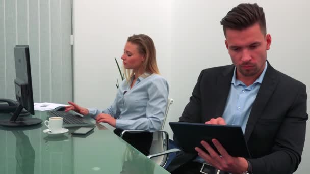 Een jonge, knappe kantoor werknemer werkt op een tablet, zijn collega werkt op een computer op de achtergrond — Stockvideo