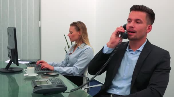 A young, handsome office worker answers the phone, his colleague works on a computer in the background — Stock Video