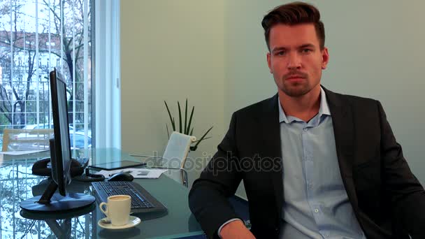 Ein junger, gutaussehender Mann sitzt an einem Tisch in einem Büro und blickt in die Kamera — Stockvideo