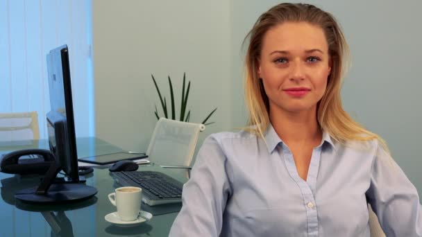 Een jonge, mooie vrouw zit op een bureau in een kantoor, knikt en glimlacht op de camera — Stockvideo