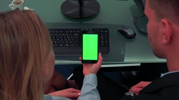 Um homem e uma mulher sentam-se em uma mesa em um escritório e olham para um smartphone com uma tela verde — Vídeo de Stock