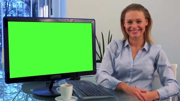 Молодая, красивая женщина сидит за столом в офисе и улыбается в камеру, зеленый экран компьютера рядом с ней повернулся к камере — стоковое видео
